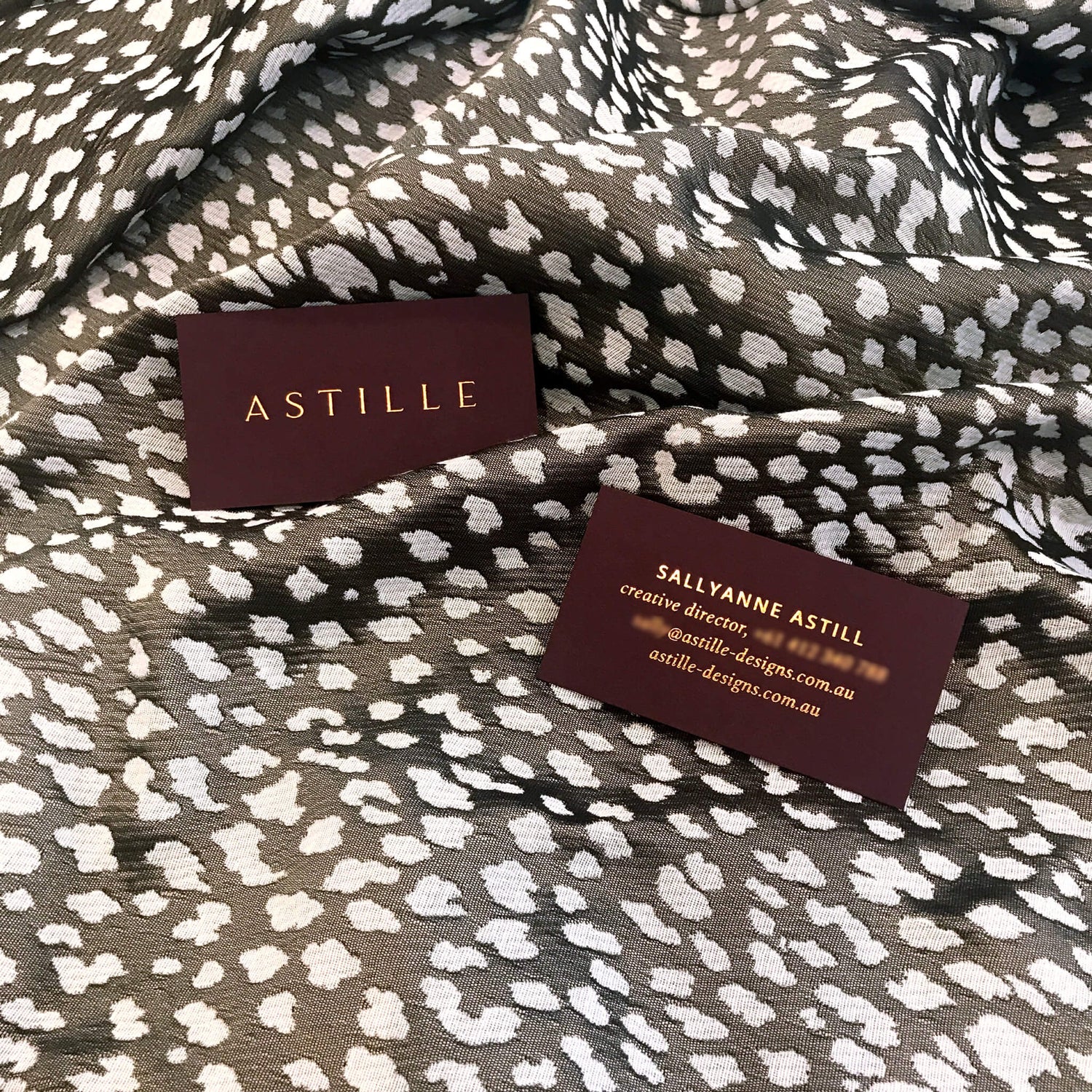 Brand designer portfolio image of the Astille fashion designer business cards on a leopard fabric background. The rich brown cardstock amplifies the copper foil business card design for an elegant, high end feel.