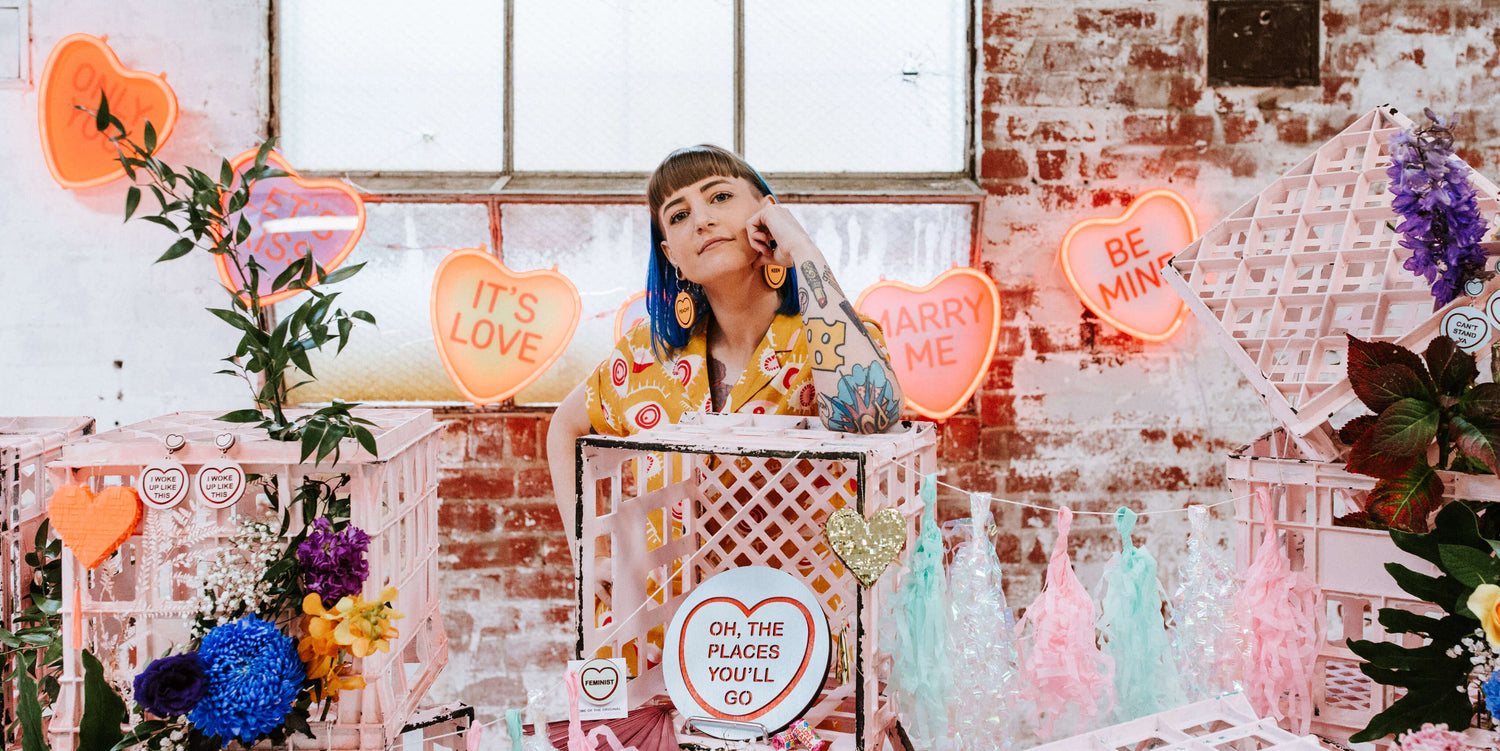 Brand identity case study image of founder Jess from Candy & Kitsch. She wears a colourful jumpsuit and stands in a sea of candy hearts and flowers. 