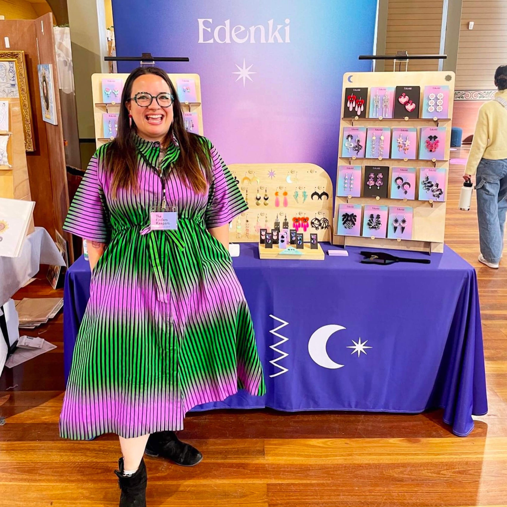 Jewellery brand identity case study image of Edenki founder Eden in front of her stall at a market. On show is a gradient backdrop with the Edenki logo in black, purple table covering with brand icon and jewellery pieces with gradient and black backing cards.