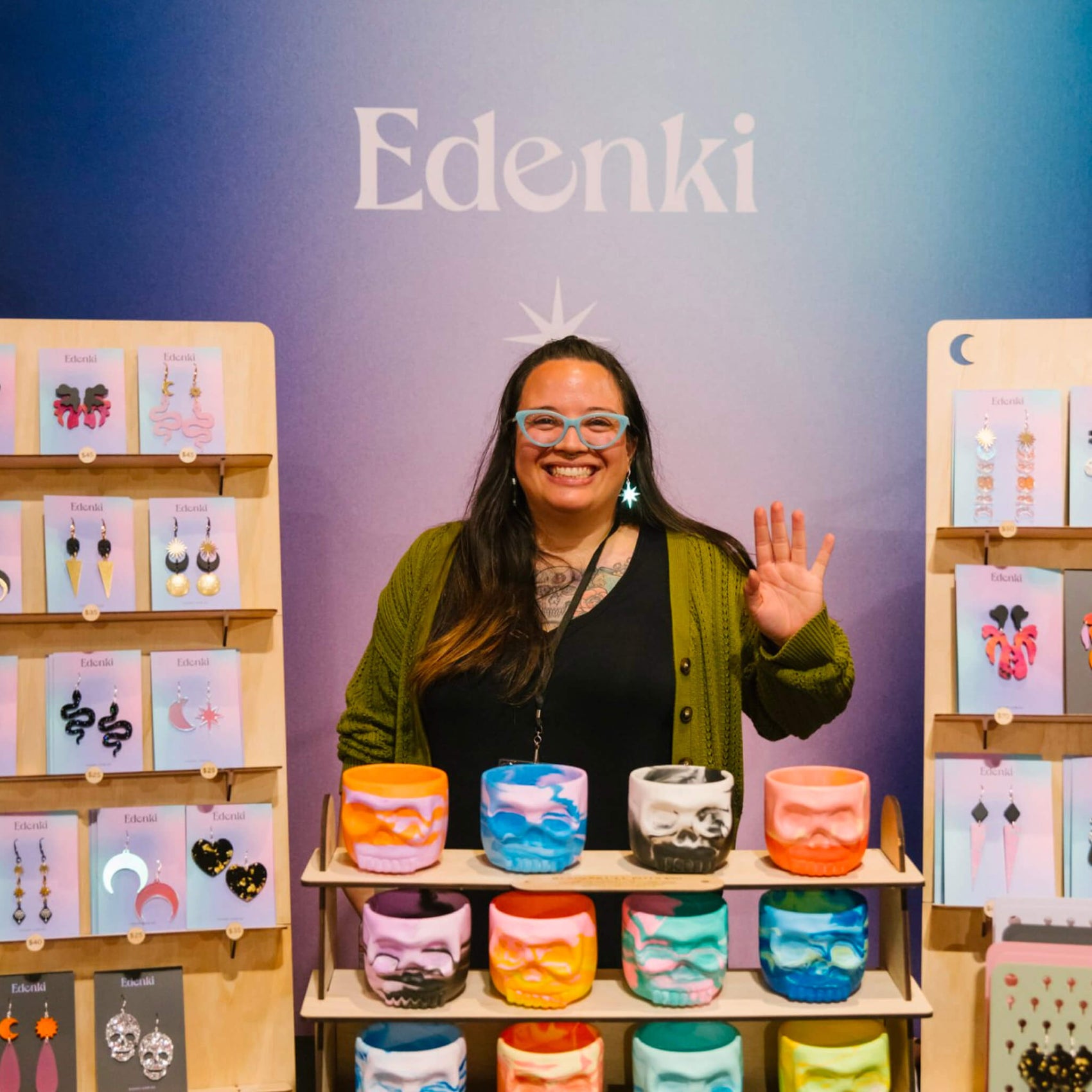 Jewellery brand identity case study image of Edenki founder Eden in front of her stall at a market. On show is a gradient backdrop featuring the Edenki logo, purple table covering with brand icon and jewellery pieces with the gradient and black backing cards.