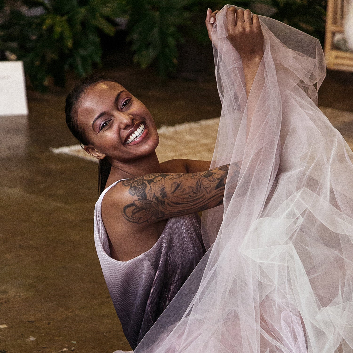 Fashion designer brand photography photograph from the Fred Archer brand identity design. A woman with arm tattoos sits on a brown floor in an ombre purple top and pink tulle skirt. 