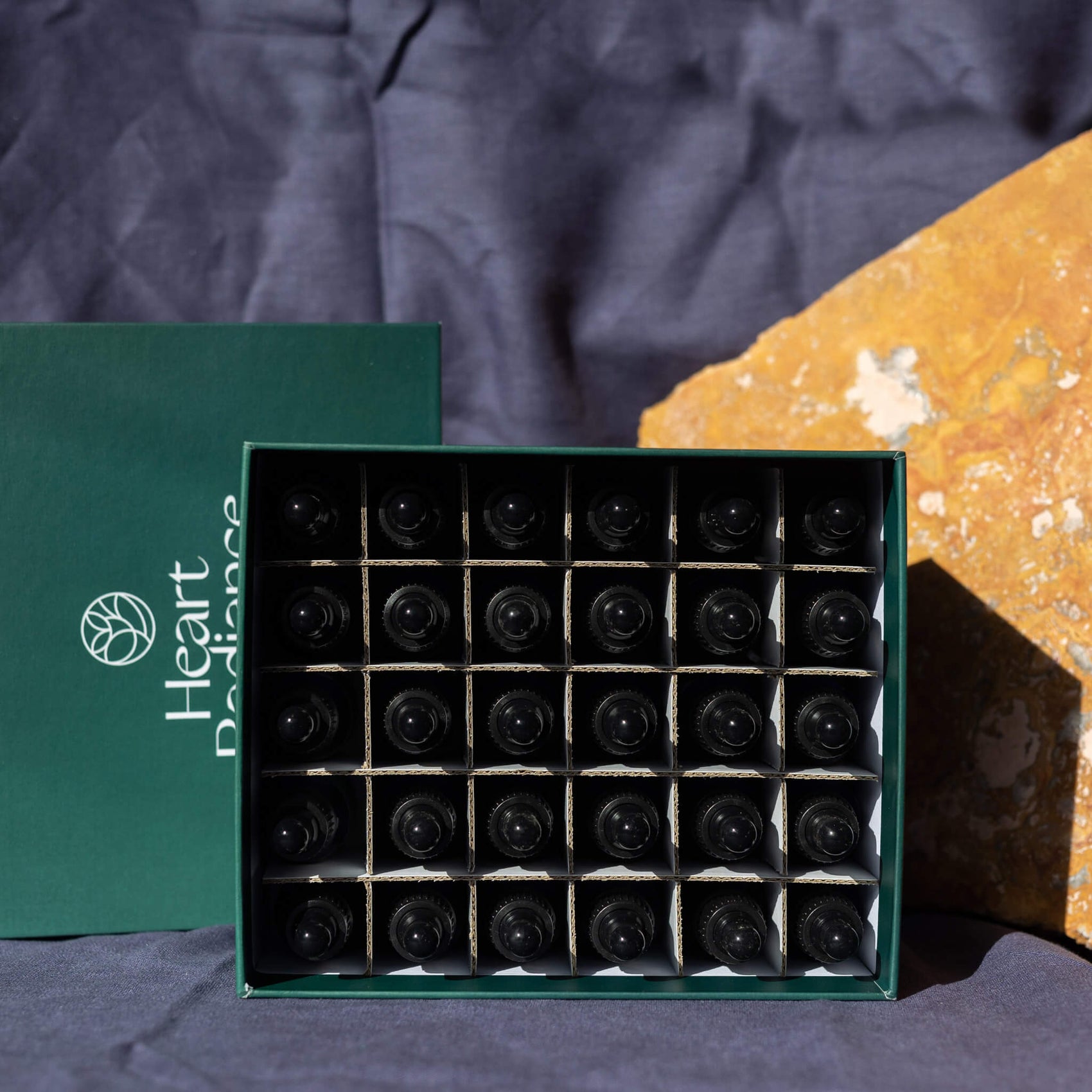 Brand identity case study image of the open HeartRadiance kit custom box design in front of stone in a deep blue background. The dropper bottles fit into individual compartments with the life featuring a deep green gradient and white logo.