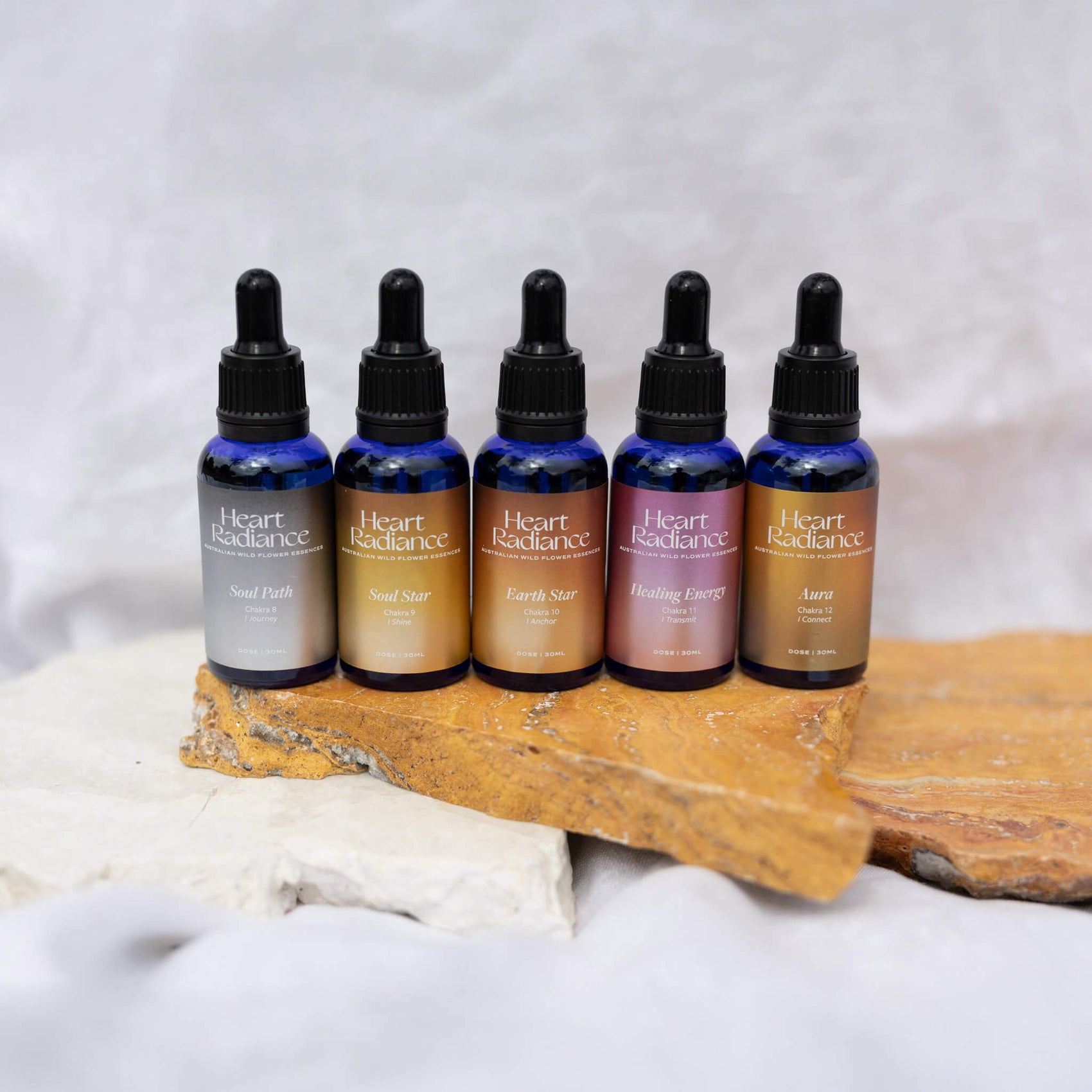 Rebranding case study image of the full range of shiny upper chakra flower essences for HeartRadiance. Each blue glass bottle features a custom designed metallic gradient (in silver, gold, copper, light pink and a mixed silver/gold/copper) with clean white typography. 