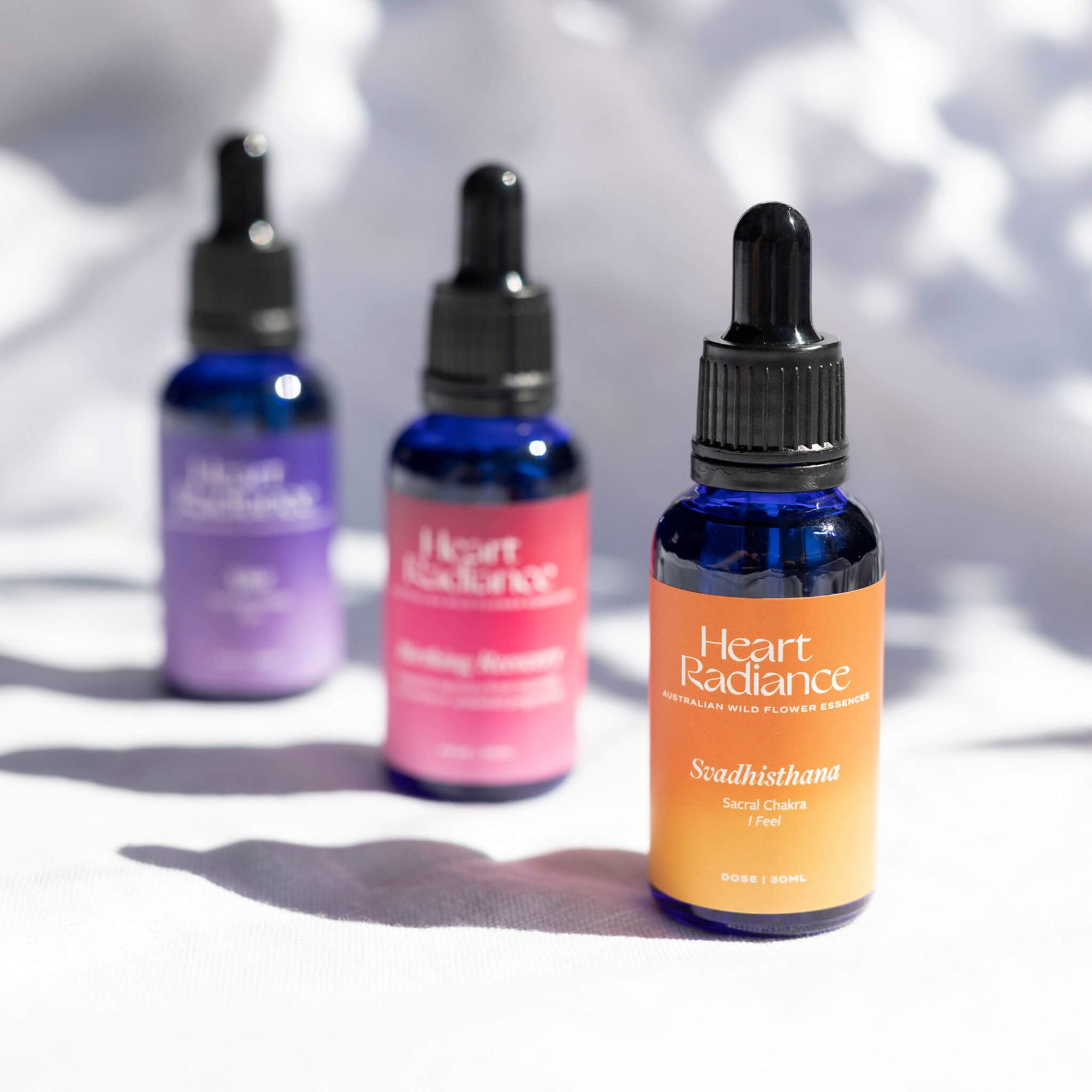 Brand identity case study image of three HeartRadiance dropper bottles in front of a white shadowy background. The bespoke product labels feature hazy gradients in purple, pink and yellow alongside elegant white typography. 