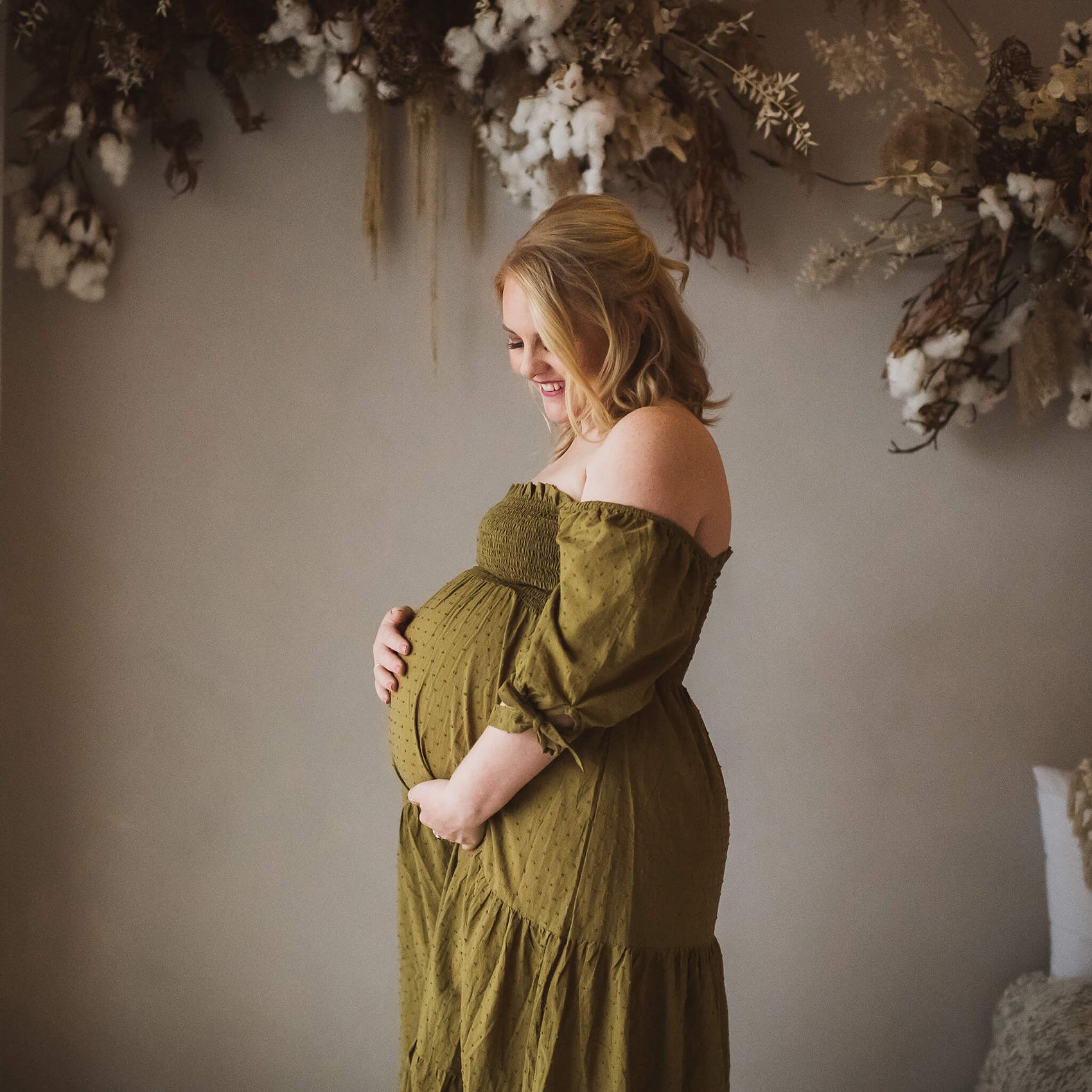 Photography logo design case study example showing imagery by Heartstory family photographers. In a neutral background surrounded by dry flowers, a blonde pregnant woman in an olive green dress holds her stomach in this maternity photoshoot image. 
