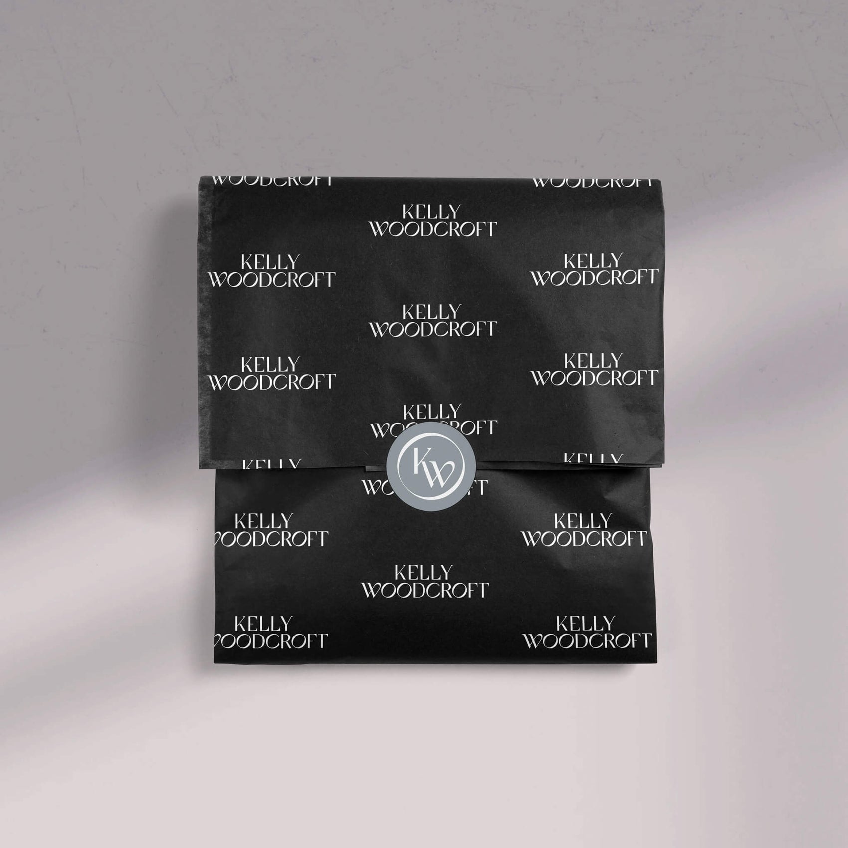 Brand design portfolio image of the Kelly Woodcroft custom tissue paper design and light grey sealing sticker. The black tissue shows the brand logo in white. 