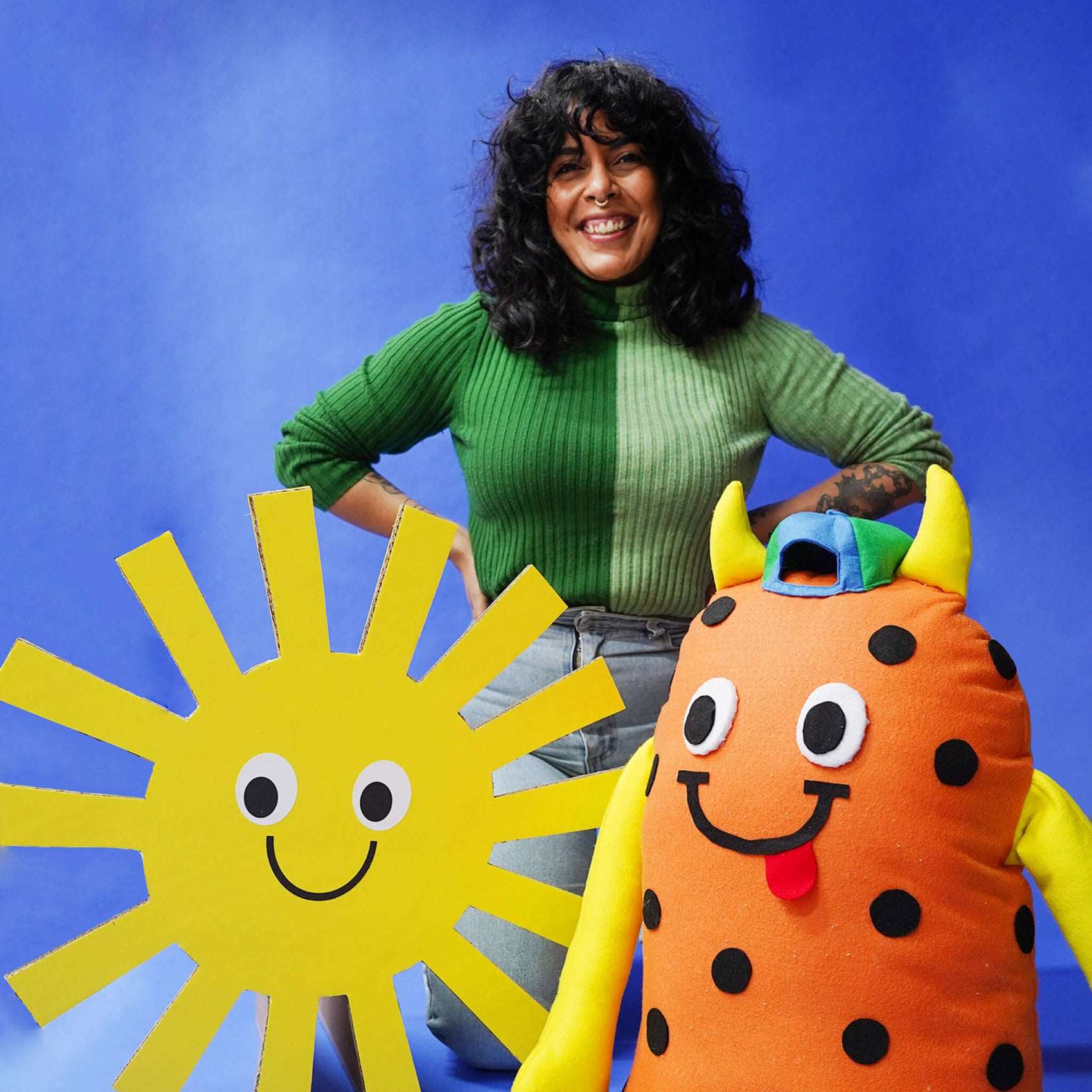 Brand designer portfolio image showing headshot brand photography of founder Kitiya. She wears a colourful green jumper in front of a blue background, two of her handcrafted props in the foreground. 