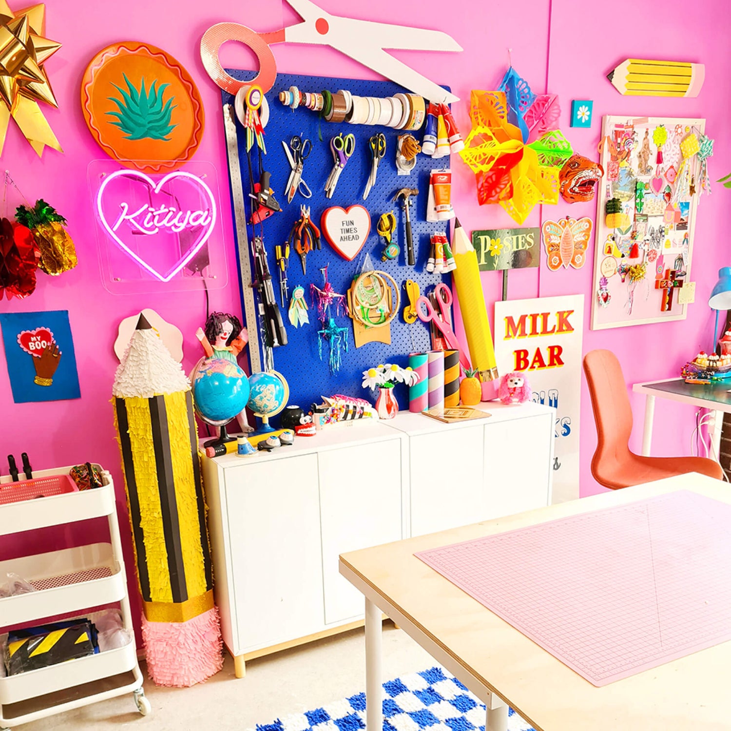 Maker brand identity case study image showing Kitiya Palaskas’ colourful studio space. From neon and vintage signs, paper ornaments, a blue pegboard and the bright pink walls, the space feels lively. 