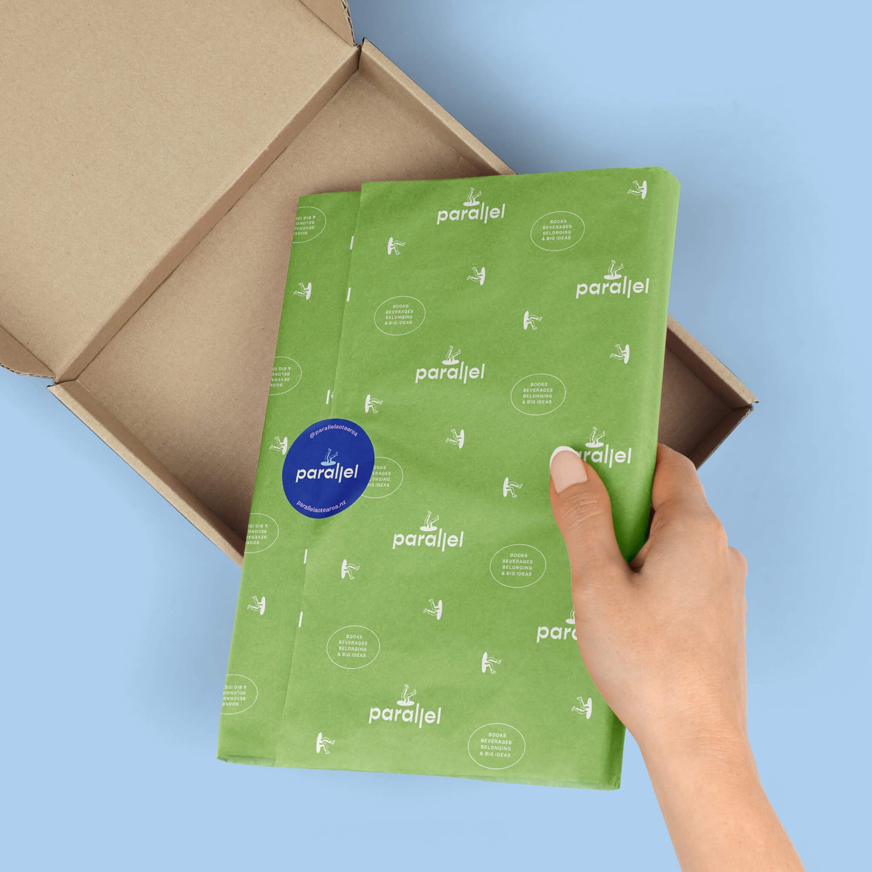Bookstore brand identity case study image of the Parallel custom tissue paper in a box. The light green design features the brand logo and supporting marks in white with a bright blue sticker with the logo sealing the package. 