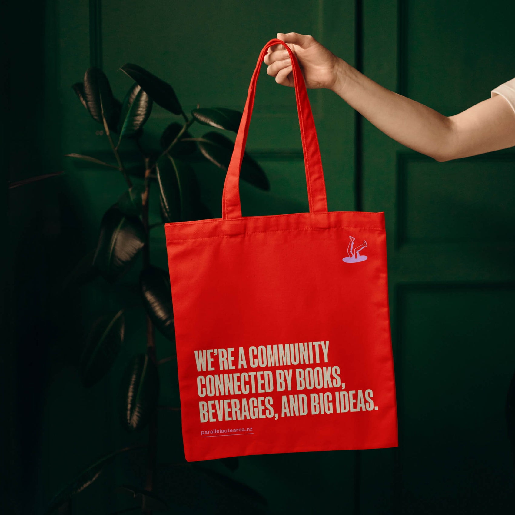 Brand designer portfolio image of a bright red Parallel tote bag. The bag features a brand tagline in pink alongside both versions of the logo and is held by an arm in a dark green room with plants.