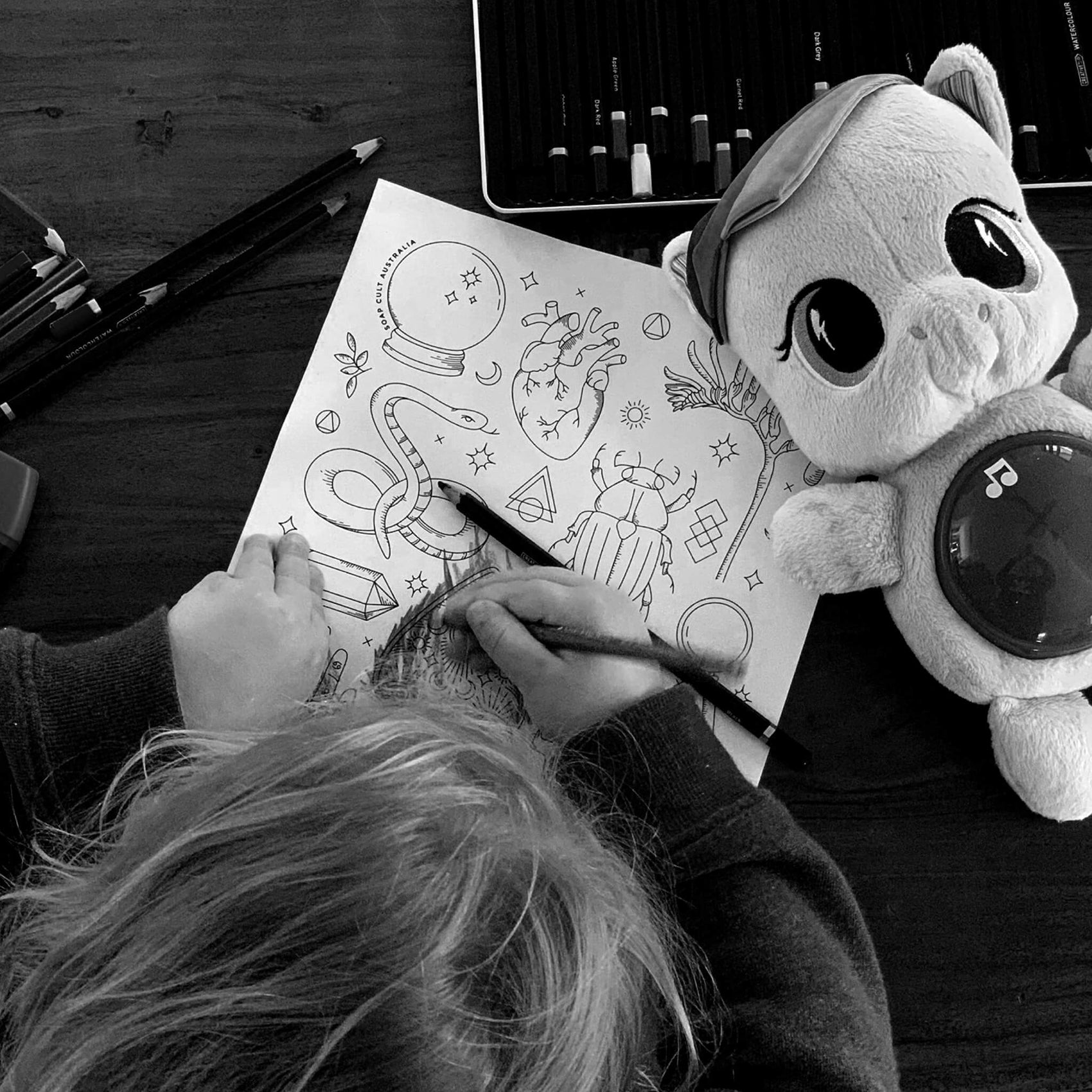 A black and white image from above of a child colouring in a Soap Cult Australia downloadable colouring in page. 