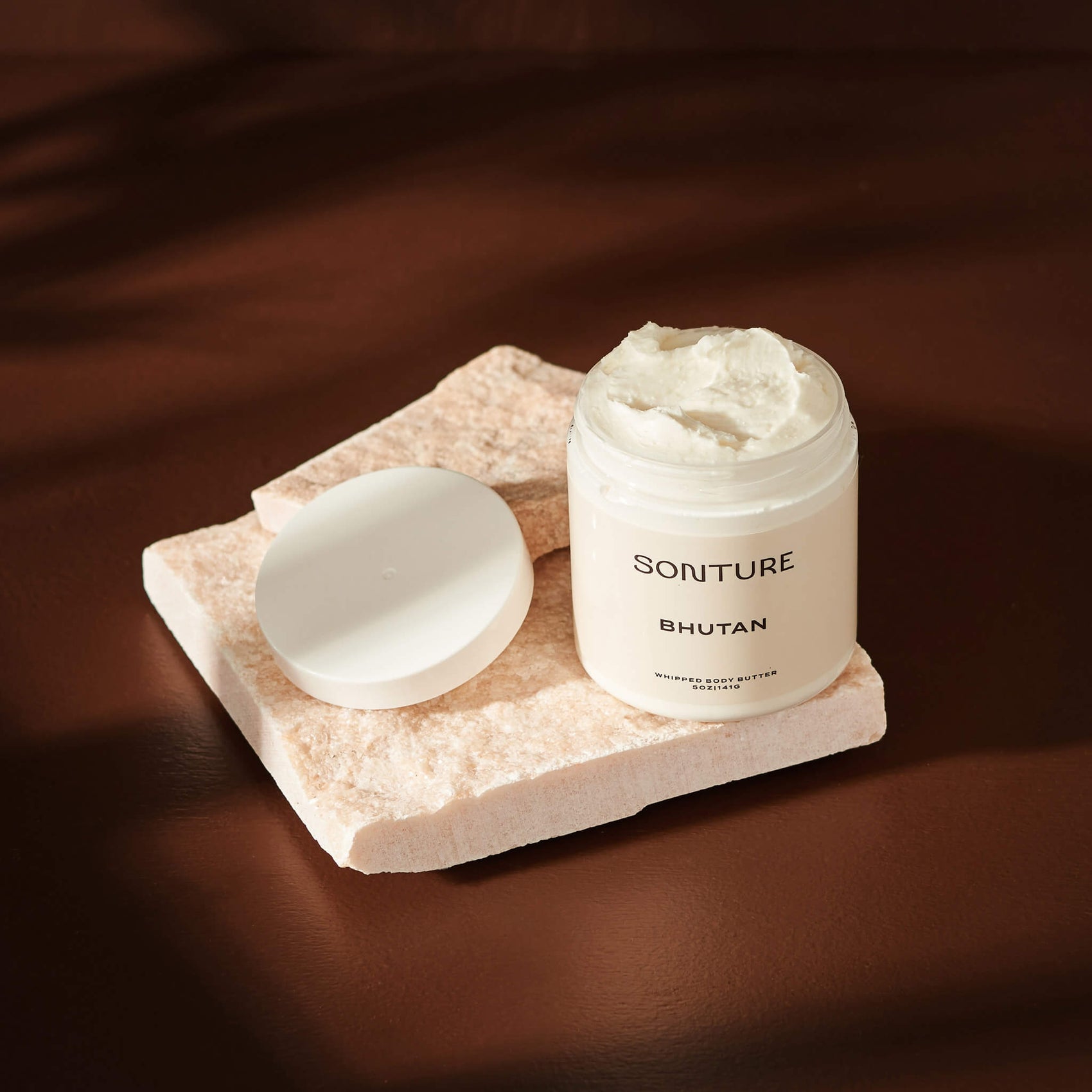 Skincare brand design portfolio image of the Sonture body scrub on a white stone in a deep brown, shadowy setting. The very minimal label creates a minimal, high end feel. 
