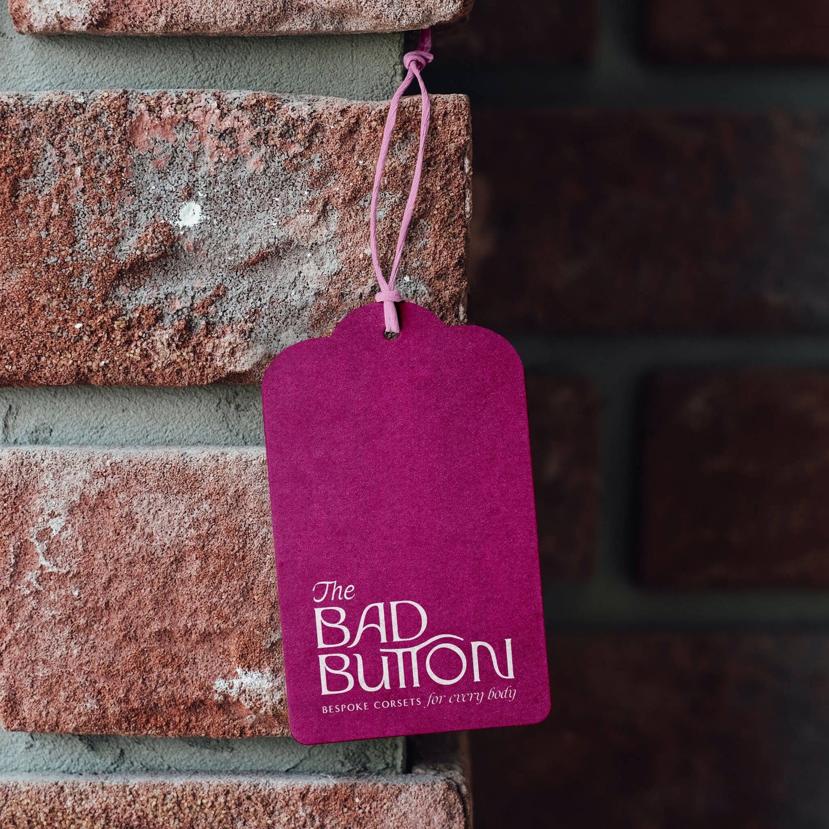 Fashion label rebranding case study image of The Bad Button swing tag on a brick wall. The tag is bright magenta and has the full brand logo on it as well as a custom-dieline of one of the arch brand support graphics. 