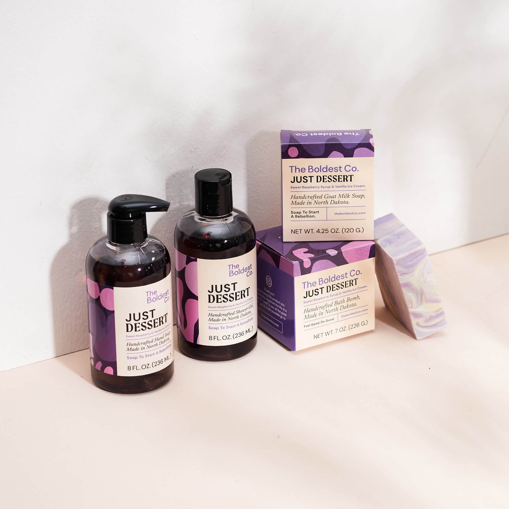 Personal care brand identity case study image of The Boldest co’s ‘Just Dessert’ scent range. A hand wash, shower gel, soap box and bath bomb box feature purple detailing and sit in a cream coloured background.