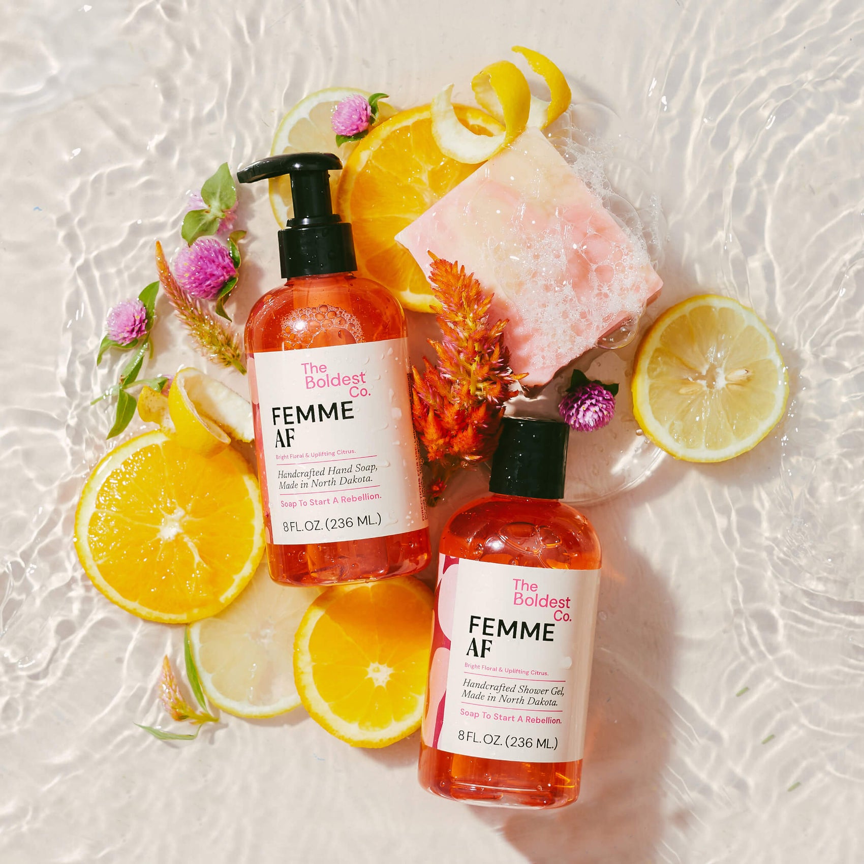 The ‘Femme AF’ product variations of The Boldest Co feature pink detailing. The products are sitting in water and are surrounded by lemons and flowers. 