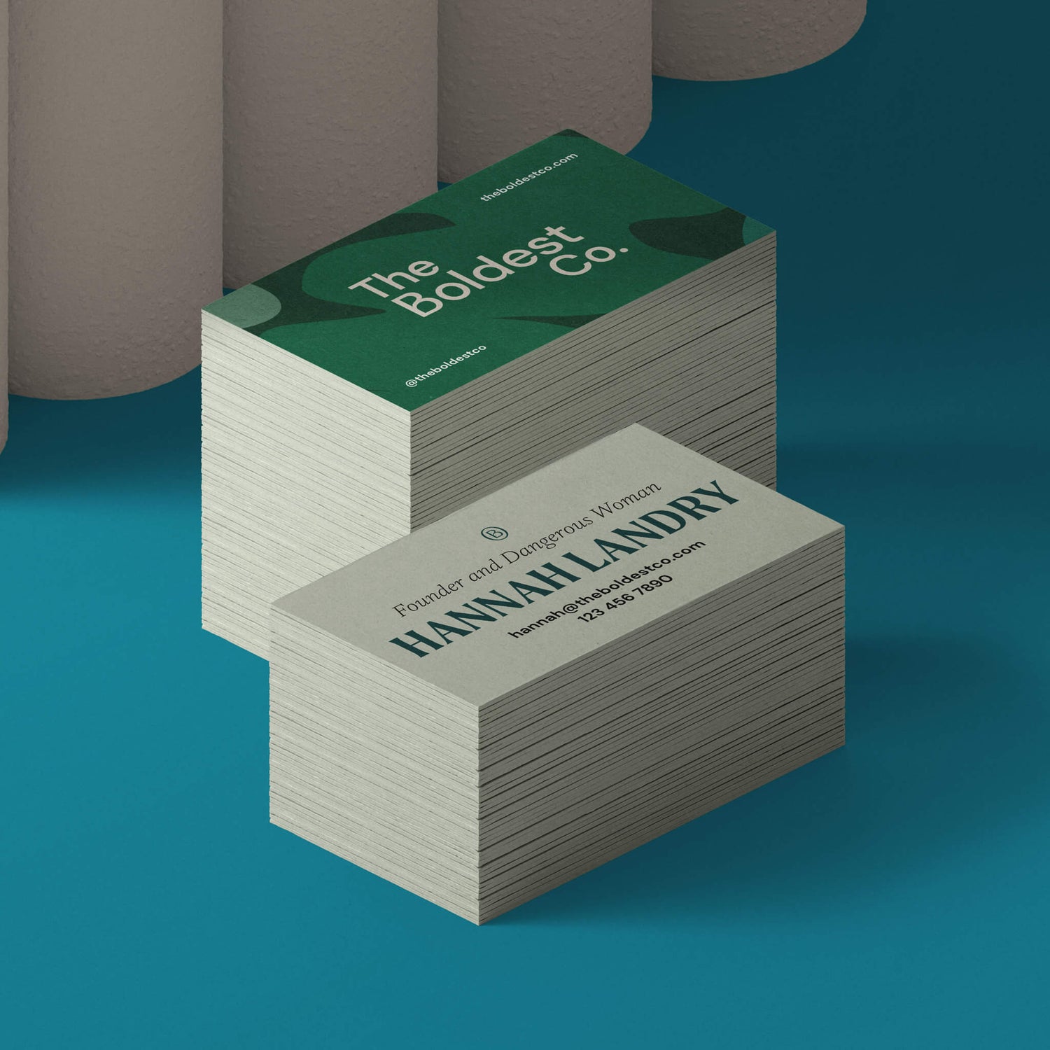 The Boldest Co business cards sit in a bright blue setting. The front of the card shows the brand logo on a green blob-textured background while the back features founder Hannah’s details and simple typesetting.