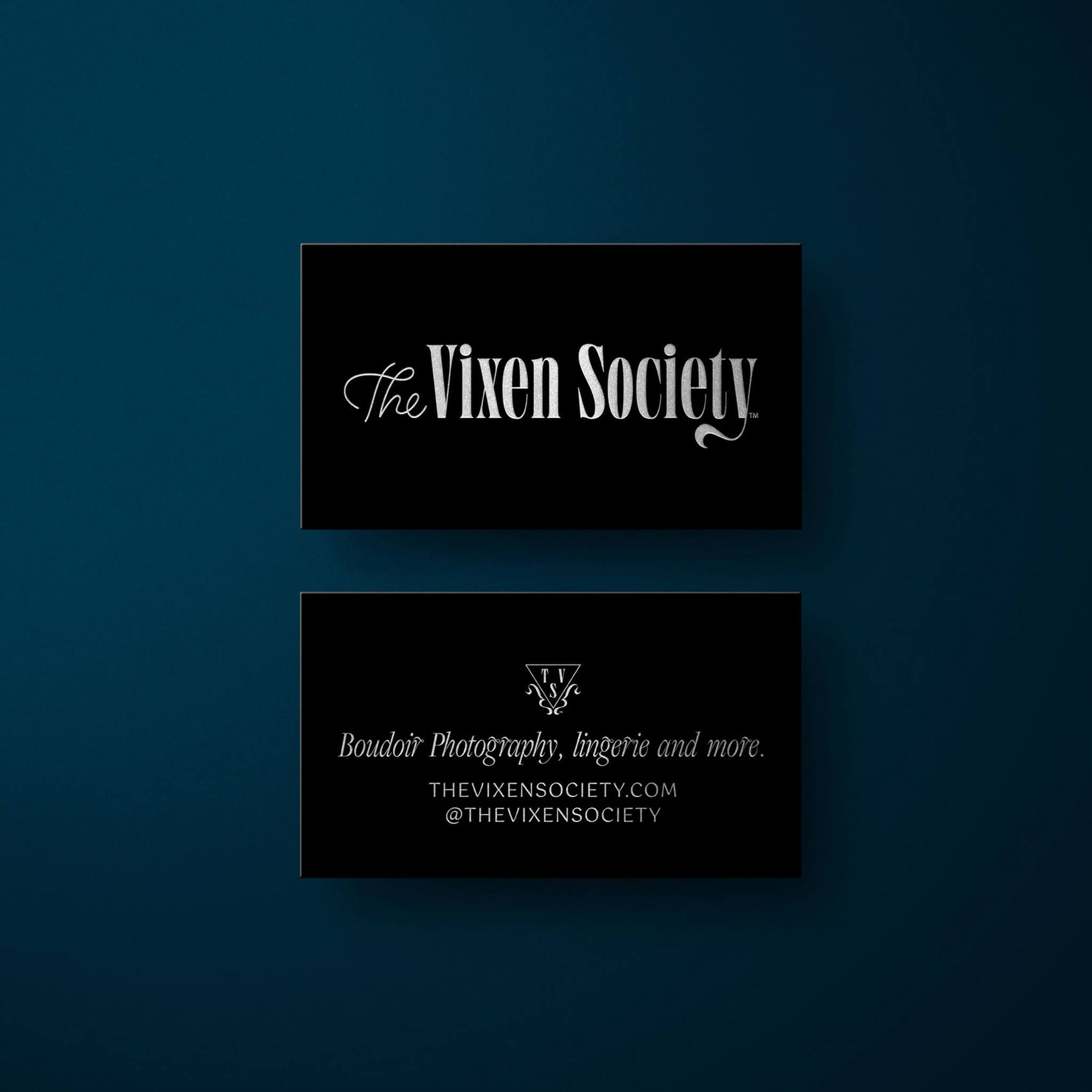 Photographer brand design examples image of the Vixen Society business cards on a deep blue background. The front features the elegant serif logo in silver foil (on black) while the back features the triangular brand icon, a tagline as well as the URL and social handles.