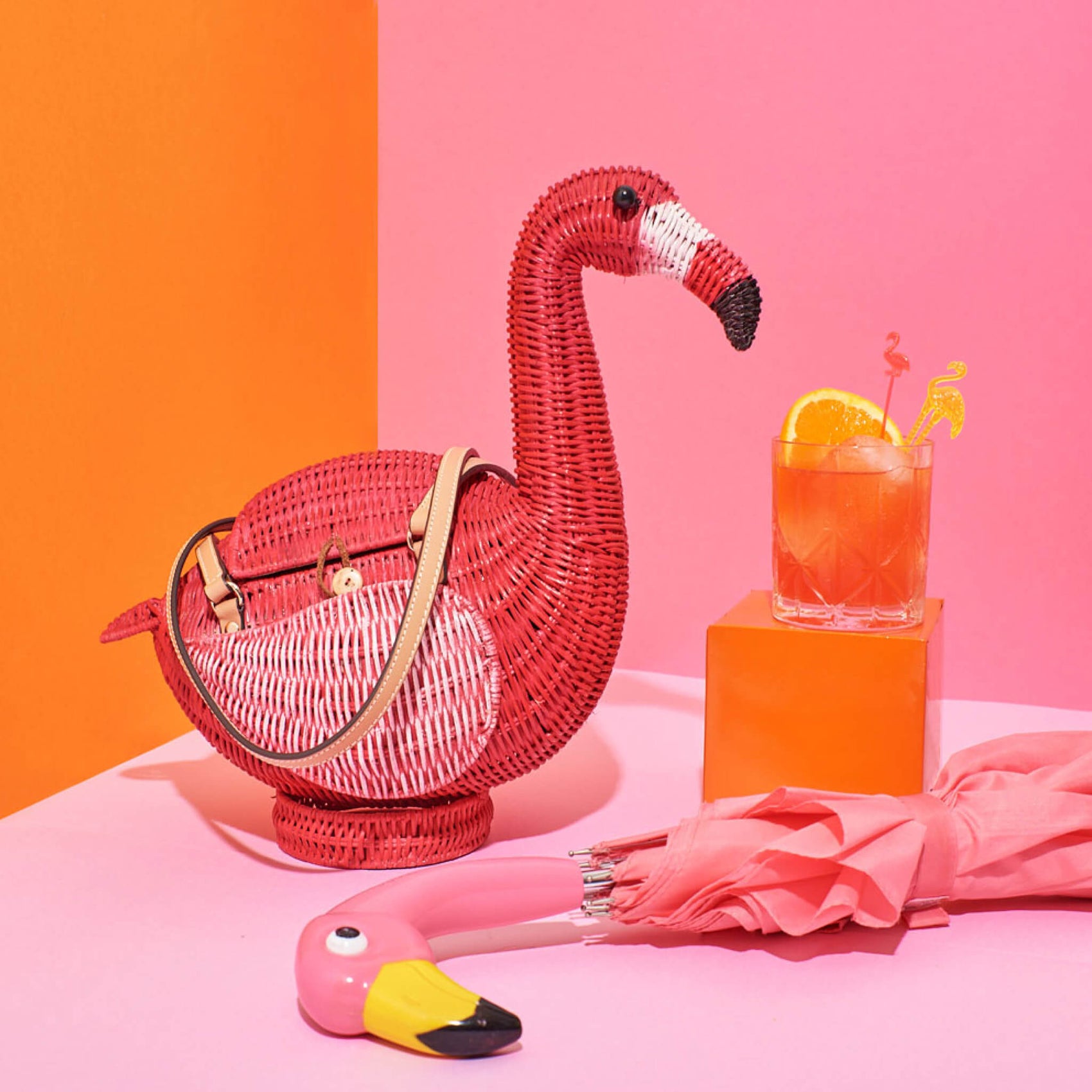 Fashion brand design example image for Wicker Darling, a high end fashion label inspired by retro palm springs. In a pink and orange background sits a bright pink wicker handbag in the shape of a flamingo, a cocktail and flamingo umbrella handle also in the shot. 