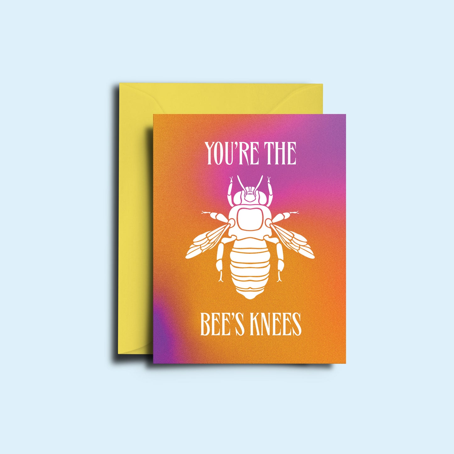 An image of the bee 'you're the bee's knees' greeting card alongside it's yellow envelope. The card features a hazy pink and orange gradient with the sharp copy and illustration of a bee in white.  