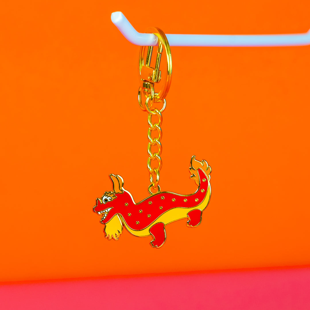 Fashion label branding design image of a Wicker Darling custom keyring design. In front of an orange background, a red and gold dragon-shaped keychain is shown.
