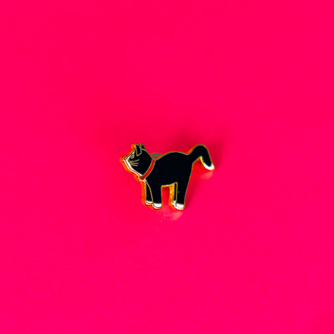 Fashion label branding design image of a Wicker Darling custom enamel pin design. In front of an pink background, a black and white cat pin with gold detailing is shown.