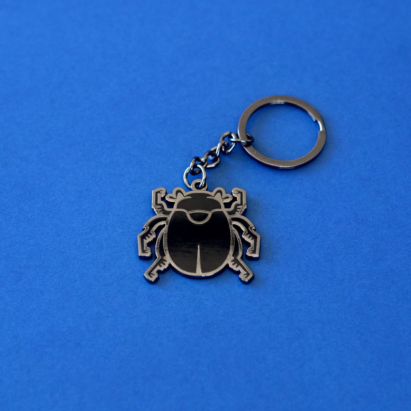On a rich blue background sits one of the Obscurio & Co. designer merch keyrings featuring the custom beetle logo icon. The keyring is a dark silver with the beetle details being in a shiny black enamel.