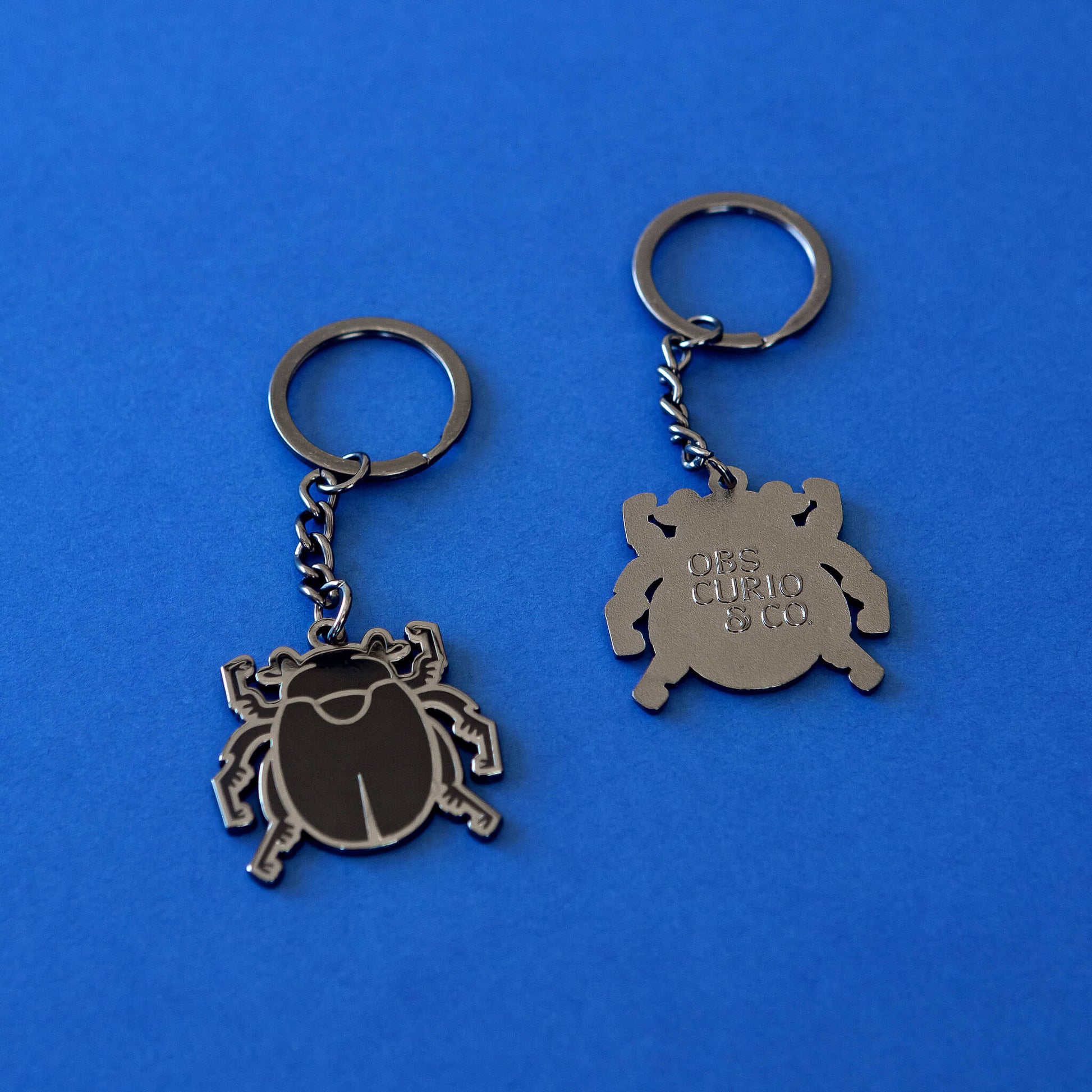 On a rich blue background sits two of the Obscurio & Co. designer merch keyrings showing the front and back of the keychain. The front of the keyring is a dark silver with the beetle details being in a shiny black enamel. The back is silver and features an embossed Obscurio & Co. logo. 