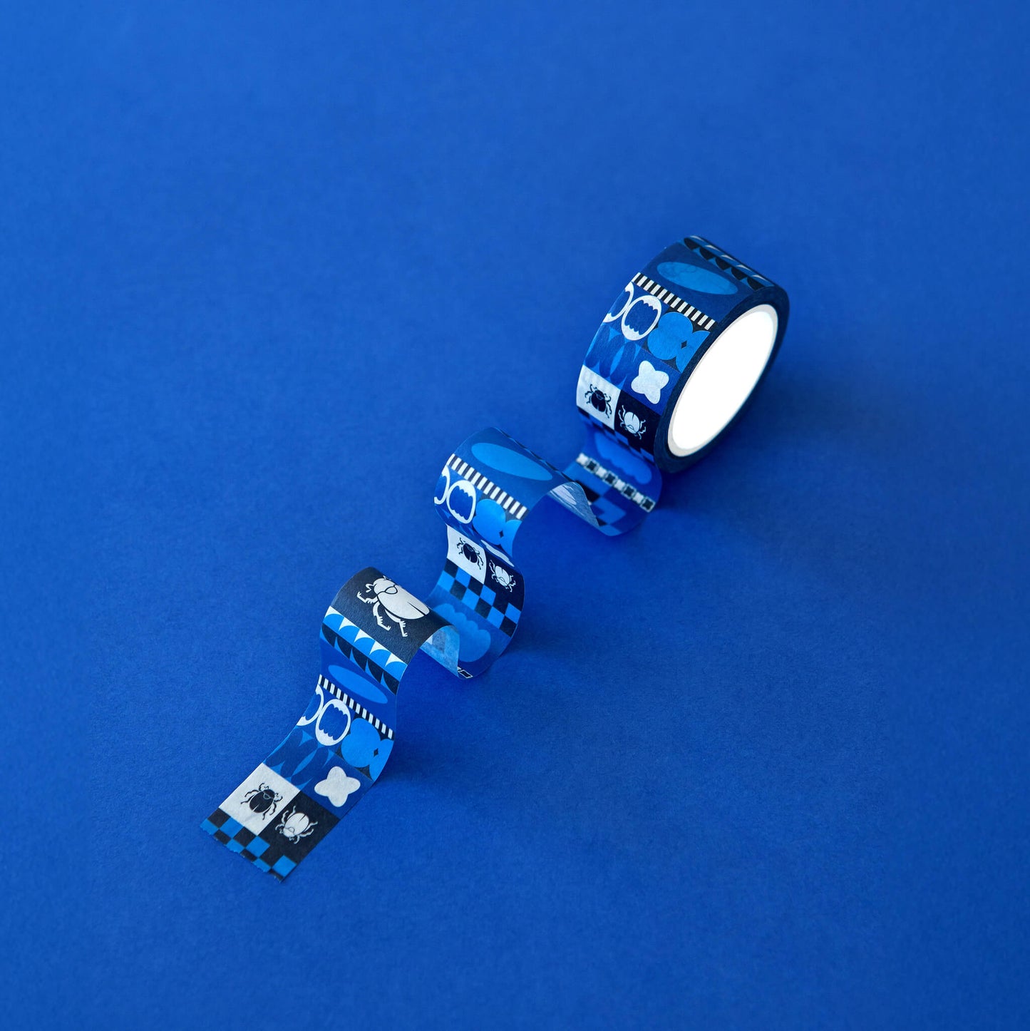 The Obscurio & Co. branded washi tape sits on a rich blue background. The custom designer washi tape features a retro inspired geometric pattern and the beetle icon in shades of several shades of blue, black and white.