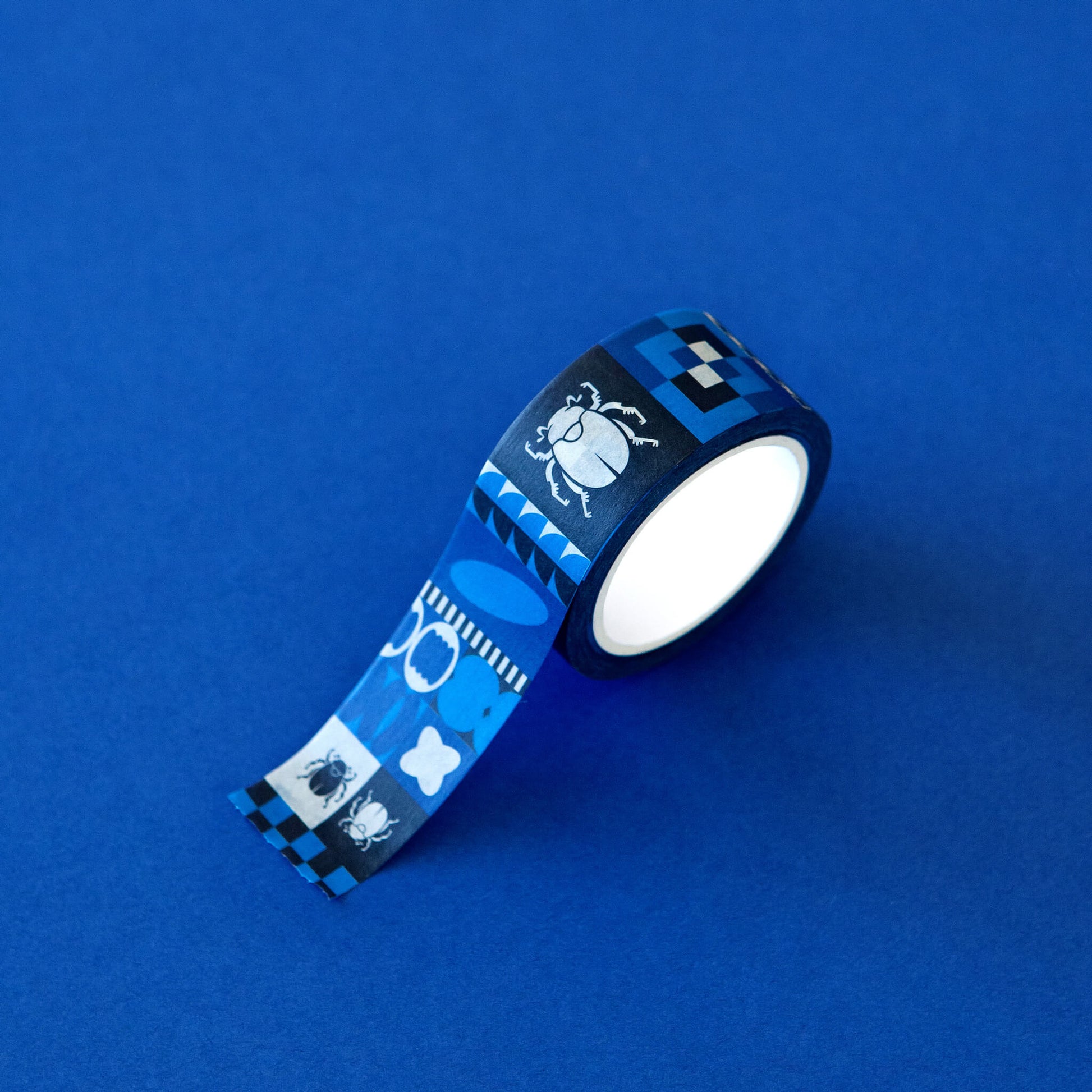 A detail image of the Obscurio & Co. branded washi tape on a rich blue background. The custom designer washi tape features a retro inspired geometric pattern and the beetle icon in shades of several shades of blue, black and white.