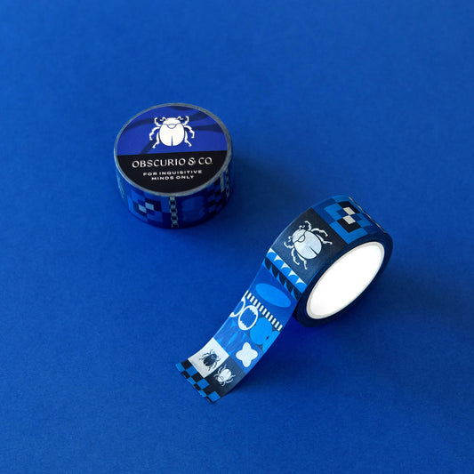 The Obscurio & Co. branded washi tape sits on a rich blue background. The custom designer washi tape features a retro inspired geometric pattern and the beetle icon in shades of several shades of blue, black and white. While one of the tapes is unrolled, the other sits with it's branded beetle sticker on it.