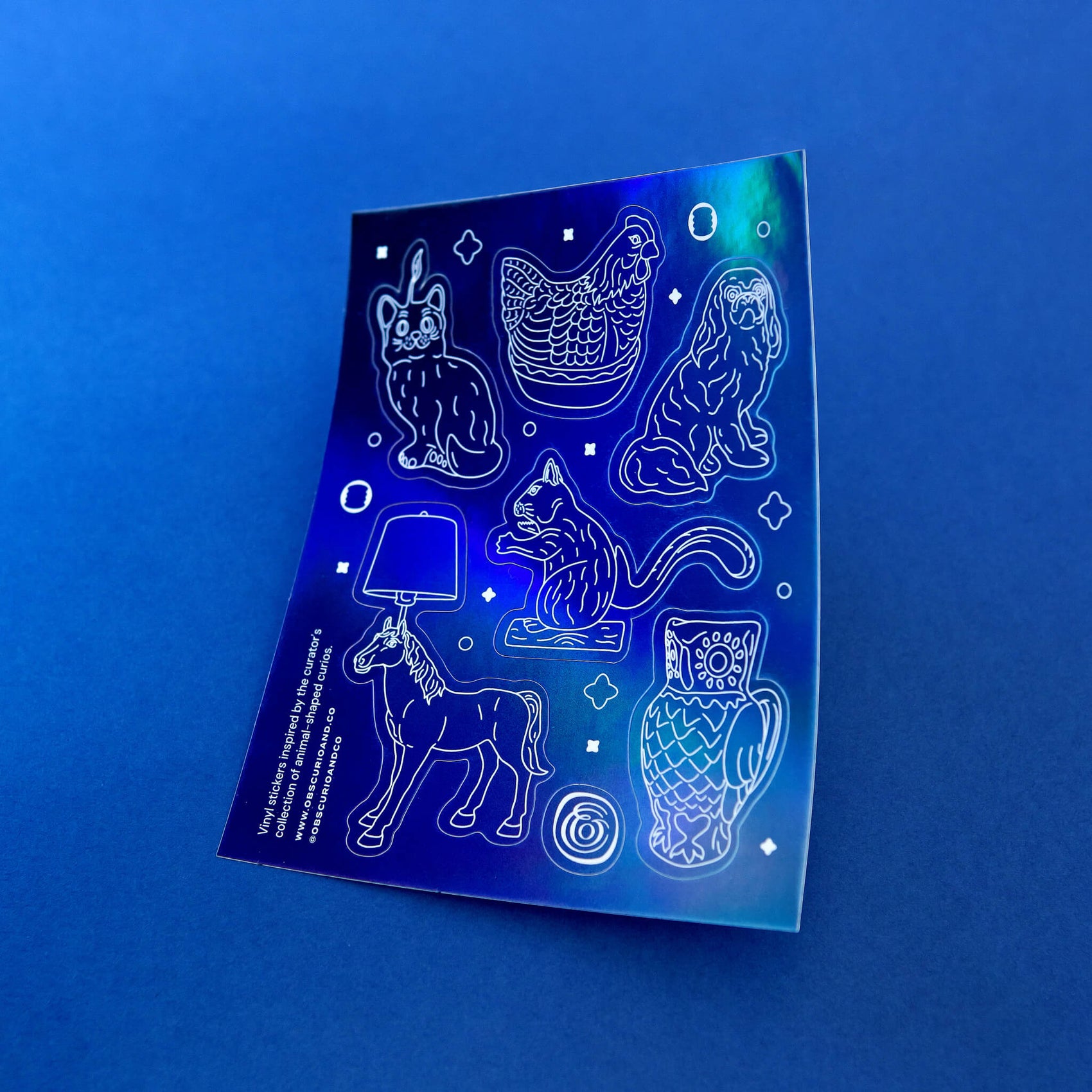 The Obscurio & Co. branded holographic sticker sheet has a deep blue subtle holographic sheet. The design features white illustrations of strange products around Laura's house like a cat candle, a horse lamp and a squirrel shaped nut cracker.